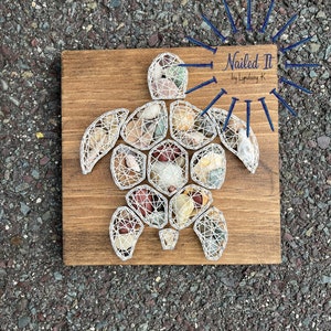 May include: A string art piece of a sea turtle made with white string and various colored stones. The turtle is on a wooden background with the text "Nailed It by Lyndsay K" in a white rectangle.