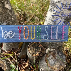 May include: A blue wooden sign with the words "be yourself" spelled out in colorful thread. The sign is mounted on a tree trunk, with the text in white, red, orange, yellow, green, and pink.