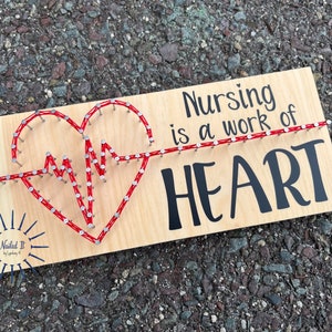 May include: A wooden sign with the text "Nursing is a work of HEART" in black lettering. A red string is wrapped around nails to create a heart shape with a heartbeat line inside.