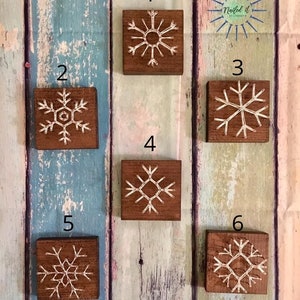 May include: Six wooden squares with white string art snowflakes on a light blue and brown wood background. The squares are numbered 1 through 6.