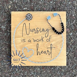 May include: A wooden sign with the text "Nursing is a work of heart" in black lettering. A stethoscope and a heart are made of string wrapped around nails. The sign is signed "Nailed It by Lyndsay K."
