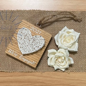 Könnte beinhalten: Ein Holz-Anhänger mit einem weißen String-Art-Herz und dem Text "2021 We're Engaged!"
