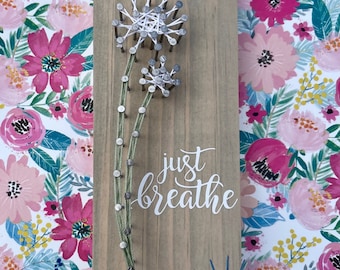 May include: String art of two white dandelions with green stems on a wood background. The text "just breathe" is written in white below the dandelions. The bottom right corner of the wood has a small logo that says "Nailed It by Lyndsey K."