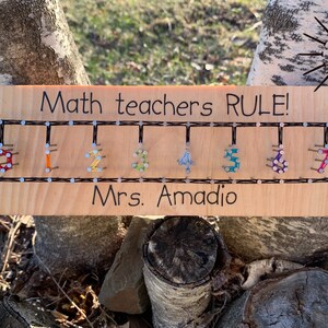 Puede incluir: Un letrero de madera con el texto "Math teachers RULE!" y los números del 1 al 7 hechos con cuerda de colores envuelta alrededor de clavos. El letrero también dice "Mrs. Amadeo" en la parte inferior.