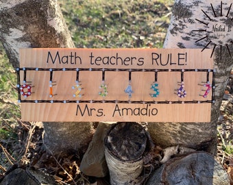 Puede incluir: Un letrero de madera con el texto "Math teachers RULE!" y los números del 1 al 7 hechos con cuerda de colores envuelta alrededor de clavos. El letrero también dice "Mrs. Amadeo" en la parte inferior.