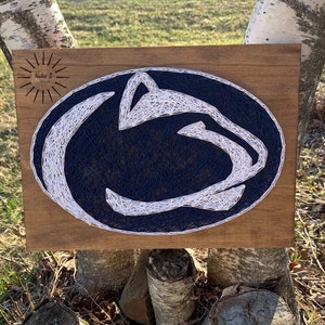 May include: A wooden sign with a blue and white string art design of the Penn State Nittany Lion mascot. The sign has a small sun with the text "Nailed It" in the top right corner.
