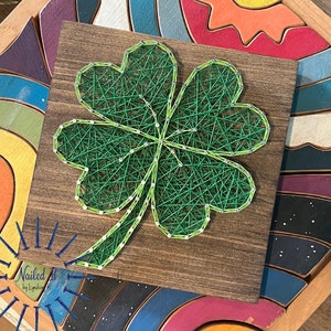 Puede incluir: Una pieza de arte de cuerda con forma de trébol de cuatro hojas de color verde sobre un fondo de madera marrón. El trébol está hecho de cuerda verde envuelta alrededor de clavos martillados en la madera. El fondo es de madera marrón oscura con veta natural. La imagen incluye el texto "Nailed It by Lyndan".