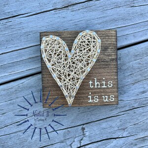 May include: A wooden sign with a string art heart and the text "this is us". The sign is on a wooden background.