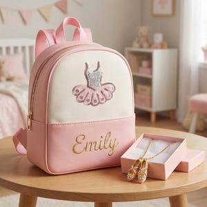 May include: Pink and white backpack with a gold zipper and straps. The backpack features a sequined ballet tutu design and the name "Emily" in gold script. A necklace with ballet shoe pendants is displayed in a pink box.