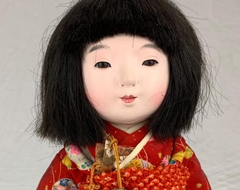 May include: A doll with black hair and a pale face wearing a red kimono featuring a pattern of colorful circular motifs and white cranes. The doll has a gold and white obi sash with a textured orange accessory and a hanging gold tassel cord.