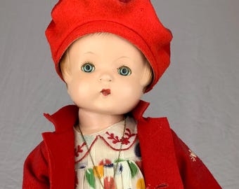 May include: A doll wearing a red felt beret and a matching red coat over a cream-colored dress with a multi-colored polka dot pattern. The doll features blue eyes, a heart-shaped gold pendant necklace with the text EFFANBEE, and red shoes.