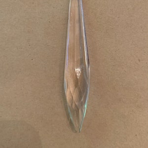 May include: A clear, faceted crystal chandelier prism with an octagonal top and a long, pointed bottom. The prism is suspended by a small, dark metal loop. The background is a neutral tan color.