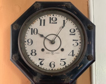 Puede incluir: Un reloj de pared antiguo de madera octogonal con una esfera blanca y marcadores de hora romanos negros. El reloj tiene una pequeña ventana redonda de vidrio con una imagen de una mujer con un sombrero. La esfera del reloj dice "MADE IN TOKYO".
