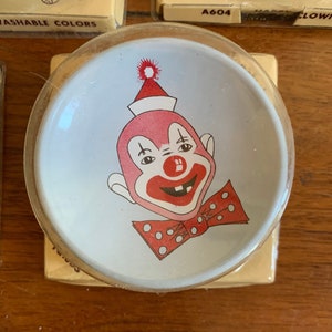 May include: A white ceramic dish with a red and white clown face design. The clown has a red nose, red lips, and a red bow tie with white polka dots. The clown is wearing a red and white hat.