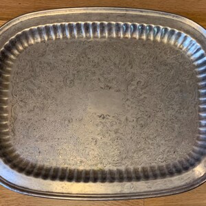 May include: Silver-toned oval serving tray with ornate handles. The tray features a detailed floral pattern across its surface and a ribbed border. The handles have a decorative, scroll-like design. The tray is displayed on a wooden surface.