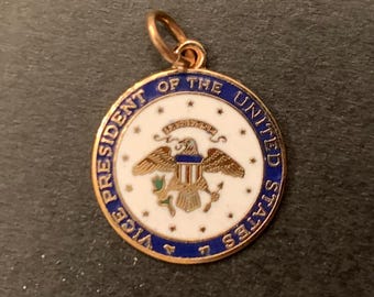May include: A circular gold-tone pendant featuring the Seal of the Vice President of the United States. The design includes a white center with a central eagle emblem, surrounded by a blue border with the text VICE PRESIDENT OF THE UNITED STATES.