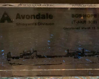 May include: A clear rectangular acrylic block featuring a black etched graphic of a ship. The text reads Avondale Shipyards Division, BOB HOPE (T-AKR 300), and Christened March 15, 1997. The block has a smooth, transparent finish with sharp edges.