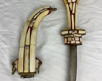 May include: A curved dagger with a handle and matching sheath featuring an intricate mosaic pattern of cream-colored segments separated by thin red lines. The handle and sheath are accented with small, circular, metallic studs and brass rings.