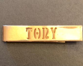 May include: A rectangular gold-toned metal tie bar featuring a polished, reflective surface. The name TONY is engraved in a stylized, serif font across the center of the bar, with the letters appearing as cutouts or deep indentations in the metal.