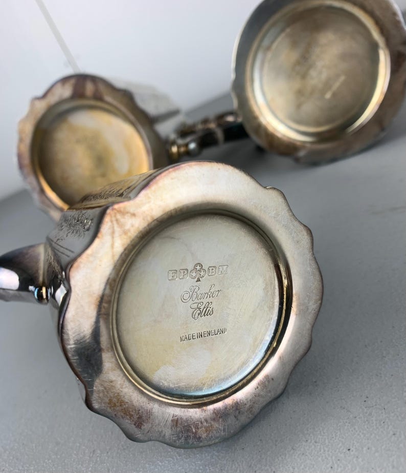 Puede incluir: Primer plano de piezas de juego de t&eacute; de plata antiguas. Las piezas tienen un dise&ntilde;o de borde festoneado y est&aacute;n estampadas con "Barker Ellis" y "Made in England". La plata tiene un aspecto deslustrado.