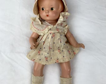 May include: A vintage doll with light brown hair, wearing a cream-colored bonnet with red trim, a matching sleeveless dress featuring a small floral and dot pattern with lace collar details, beige ribbed socks, and tan buckled shoes.