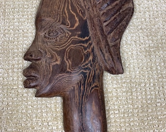 May include: A dark brown wooden wall carving depicting the profile of a human head. The sculpture features visible wood grain patterns throughout the surface, with a stylized hair design and defined facial features including eyes, nose, and lips.