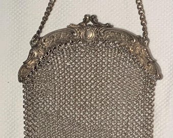 May include: A silver-toned metal mesh purse featuring a decorative, ornate metal frame with intricate scrollwork and a central circular detail. The bag includes a matching chain handle and five small, spherical metal charms hanging from the bottom edge.