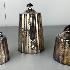 Puede incluir: Un juego de t&eacute; plateado con tetera, azucarero y lechera. El juego tiene un dise&ntilde;o acanalado y detalles ornamentados. La tetera tiene un adorno negro en la tapa. La lechera y el azucarero tienen asas.