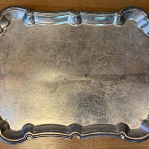 May include: A rectangular silver-colored serving tray with ornate handles. The tray has a decorative, scalloped edge and a slightly textured surface. The handles feature a detailed, floral design. The tray is likely vintage or antique.