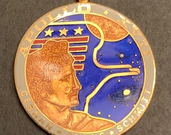May include: A circular gold-toned metal pendant featuring a colorful enamel design of the Apollo XVII mission patch. It depicts a profile of the Greek god Apollo, three stars, a red striped flag, a planet, and space imagery with the text APOLLO XVII.