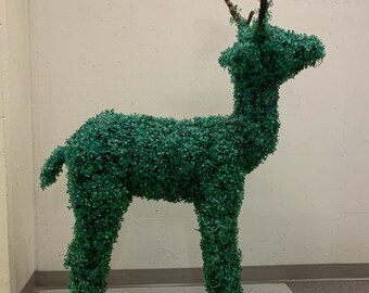 May include: A standing deer sculpture covered in dense, dark green artificial foliage. The figure features a textured, leafy exterior, a short tail, and prominent brown, branch-like antlers extending from the top of the head.