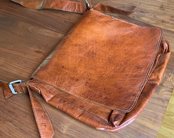 May include: A tan leather messenger bag featuring a large front flap, a long adjustable shoulder strap with a silver-toned metal buckle, and visible stitching along the edges. The leather has a textured, worn appearance with natural variations.