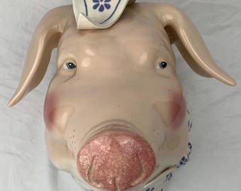 May include: A ceramic pig figurine featuring a glossy light pink finish with rosy cheeks and a textured pink snout. The pig wears a white neckerchief and a matching headpiece, both decorated with a repeating pattern of small blue flowers.