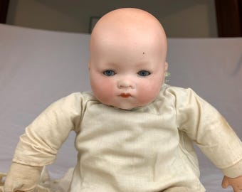 May include: A doll with a smooth, pale, bald head, blue eyes, and rosy cheeks. The doll has a small, defined nose and painted red lips. It is dressed in a long-sleeved, off-white, plain fabric garment that covers the torso and arms.