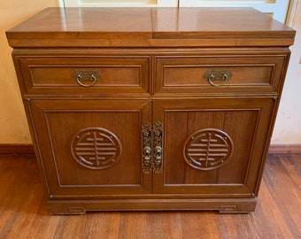 Puede incluir: Un mueble de madera marrón medio con dos cajones y dos puertas. Tiene herrajes de latón y un diseño circular tallado en las puertas. Este mueble probablemente se utiliza para el almacenamiento.