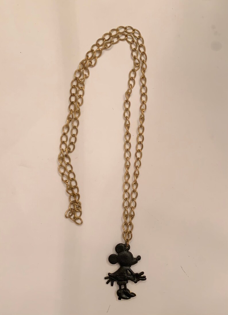 Puede incluir: Un collar de cadena dorada con una silueta negra de Minnie Mouse como colgante.