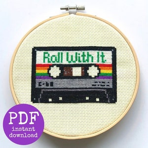 Puede incluir: Un patrón de punto de cruz de una cinta de casete con el texto "Roll With It" en él. La cinta de casete está rodeada por un borde de arcoíris. El patrón está en un aro de bordado de madera.