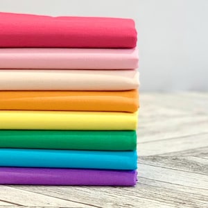 May include: A stack of eight folded fabric pieces in a spectrum of colours, including hot pink, light pink, cream, orange, yellow, green, bright blue, and purple. Each piece of fabric is neatly layered on top of the other in a vertical column.