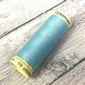 May include: A spool of light blue Gütermann thread. The cylindrical spool has a light yellow base and top. The text "Gütermann" is printed on the top. The spool contains approximately 100 metres of thread.