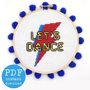 May include: A white embroidery hoop with a blue pom-pom trim. The hoop contains a cross-stitch design of a red and blue lightning bolt with the text "LET'S DANCE" in yellow letters.