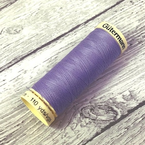 May include: A spool of lavender-coloured Gütermann thread. The spool is cylindrical with a cream-coloured label that reads "Gütermann" and "110 yds/vgs". The thread is tightly wound and appears smooth. The background is a light-coloured wooden surface.
