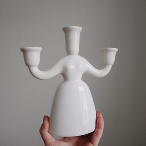 Könnte beinhalten: Ein weißer Kerzenhalter aus Keramik mit drei Kerzenschalen. Der Halter hat eine stilisierte menschliche Form, mit einer Mittelsäule und zwei Armen, die sich ausstrecken, um die Kerzenschalen zu halten. Der Sockel ist ausgestellt. Der Halter wird von einer Hand gehalten.