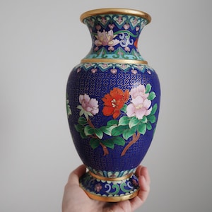 May include: A blue decorative vase with a floral design. The vase features a gold rim and base, with a detailed pattern of flowers in shades of pink, red, and white. The vase is held by a person.