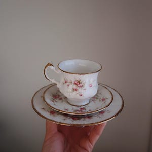 Könnte beinhalten: Ein weißes Teeservice mit rosa Rosenmuster und goldenem Rand. Das Set besteht aus einer Tasse, einer Untertasse und einem Teller. Die Tasse hat einen geschwungenen Henkel und steht auf der Untertasse und dem Teller.