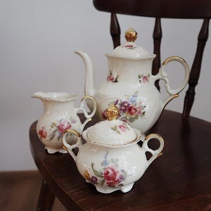 Puede incluir: Un juego de té de porcelana vintage con tetera, jarra de crema y azucarero. El juego es blanco con ribetes dorados y un estampado floral con rosas rosas, moradas y azules. El juego está sobre una silla de madera.