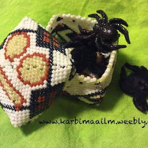 Peut inclure: Une petite boîte blanche en perles avec un motif géométrique noir, rouge et jaune. La boîte est ouverte et une araignée noire scintillante est à l'intérieur. Une autre araignée noire scintillante se trouve sur la surface en tissu vert à côté de la boîte. Le texte "www.karbimaailm.weebly.com" est visible sur le tissu.