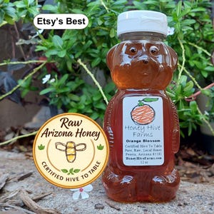 May include: A bear-shaped bottle filled with orange-colored honey. The label reads "Honey Hive Farms Orange Blossom" and "Raw Arizona Honey". The bottle is 12 oz. and has a white cap. The background is green foliage.