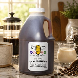 May include: A large, dark-colored plastic jug with a white cap and handle. The jug has a label with a bee graphic and the text "Honey Hive Farms LOCAL WILDFLOWER". The jug contains a dark liquid, likely honey, and is labeled as 6 lbs.