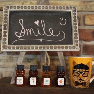 May include: A chalkboard sign with the word "Smile" in pink script, a heart, and a smiley face. Four bear-shaped honey bottles and a yellow mug with a cat wearing glasses and a bow tie are in front of the sign.