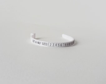 Pode incluir: Uma fita medidora de anéis de plástico branco com texto impresso em preto que diz SIZE & HALF SIZES seguido de uma escala numérica de 1 a 13 com marcas para meios tamanhos, projetada para medir a circunferência do dedo.